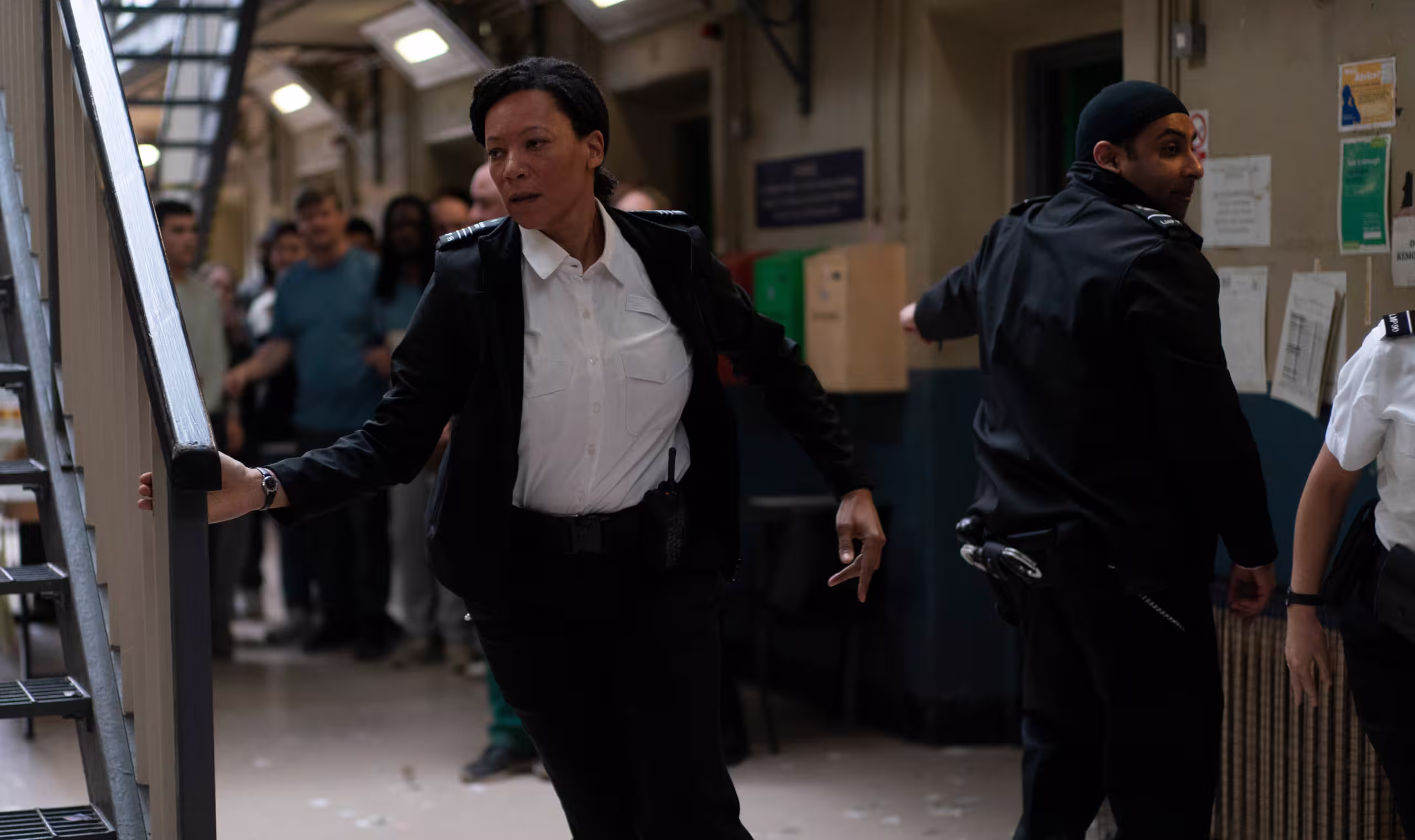 Female officer running through a busy cell block as other guards restrain prisoners.