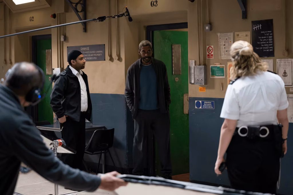 Man and woman prison officers speaking to a convict in a cell block, with a production crew member visible holding a light reflector in the background.
